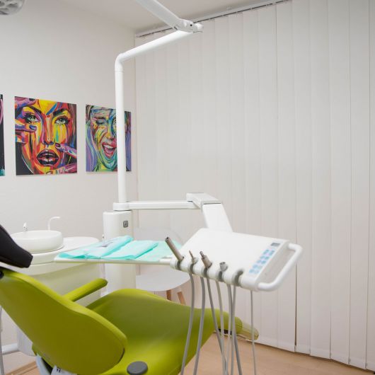 Behandlungsraum mit einem grünen Zahnarztstuhl, verschiedenen zahnärztlichen Instrumenten und bunten abstrakten Porträts von Gesichtern an der Wand. Vertikale weiße Jalousien verdecken das Fenster.