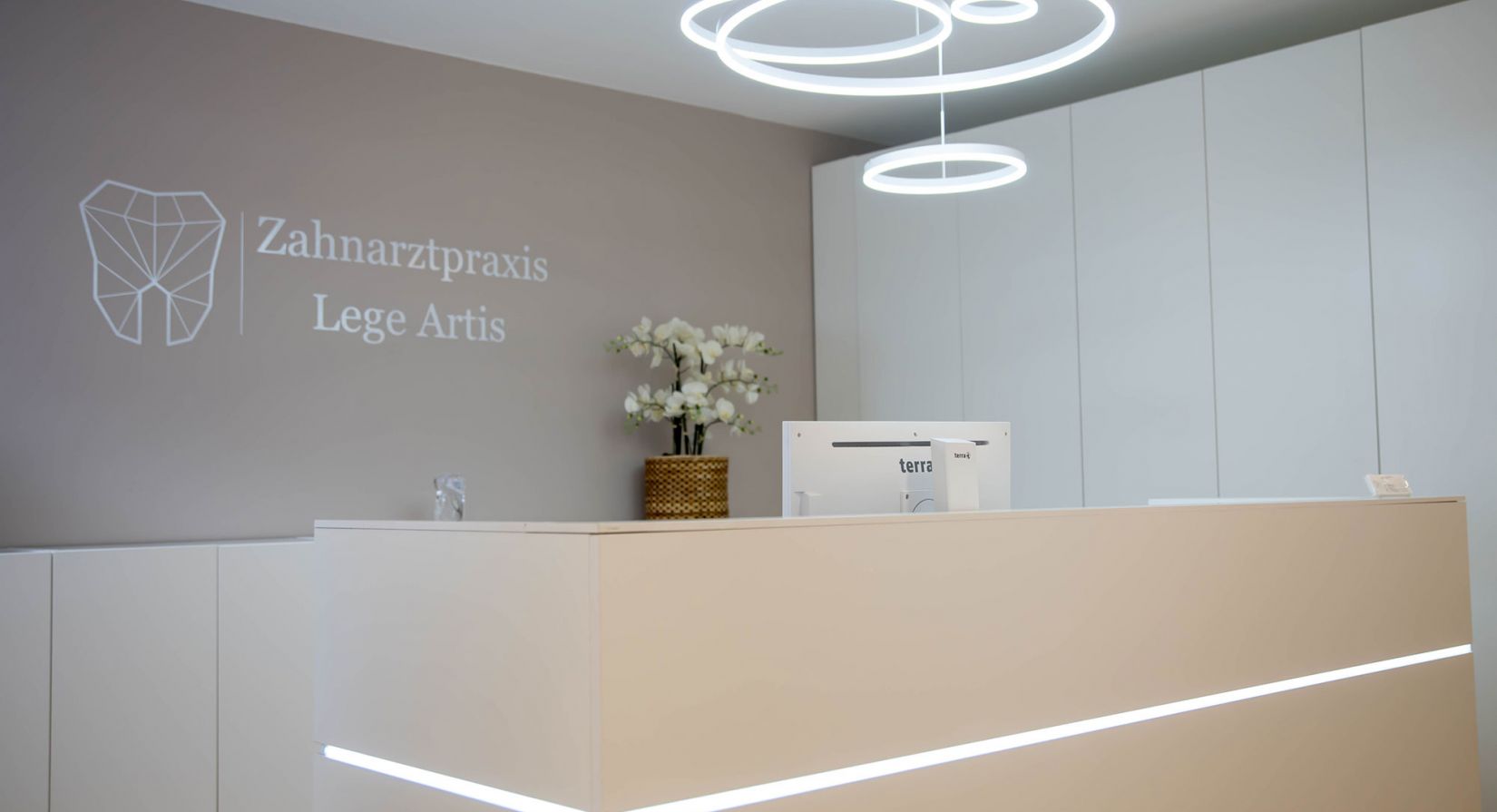 Moderner Empfangstresen mit einem schlichten weißen Schreibtisch, minimalistischem Dekor, einem Computermonitor und weißen Schränken. Die Wand zeigt das Logo der Zahnarztpraxis Lege Artis mit einem geometrisches Zahnlogo.